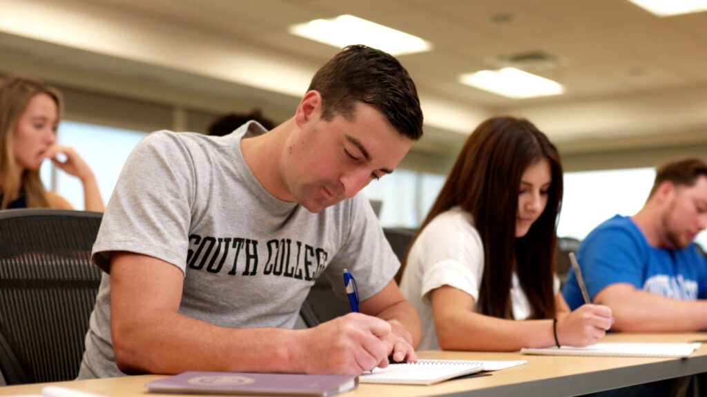 Four students are seated in a classroom, focused on writing in their notebooks. The student in the foreground is wearing a "South College" t-shirt.