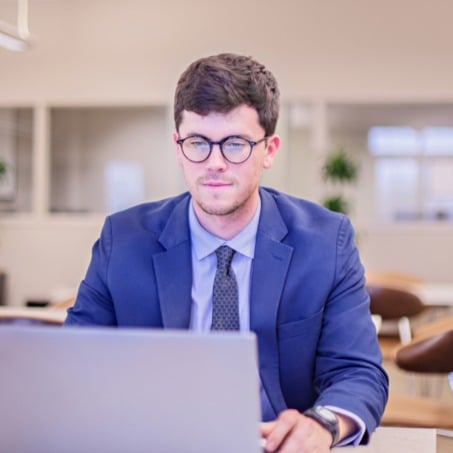 A male business student in a blue suit, glasses, and a tie is sitting and working on a laptop. The background features a modern office setting with plants and bright lighting.