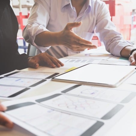 Two people in a meeting room engaged in discussion. One person gestures towards a table covered with diagrams, charts, and a tablet. Both are wearing professional attire, and the atmosphere suggests a collaborative work session, possibly related to project planning.