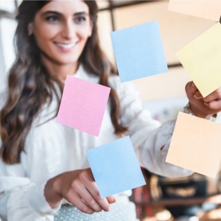 A student teacher with long brown hair wearing a white blouse is smiling while placing colorful sticky notes (blue, pink, peach, and yellow) on a glass surface. The background appears to be a modern office environment.
