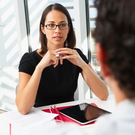 A legal studies student with glasses, wearing a black dress, sits at a desk with a tablet and documents in front of her. She appears to be in conversation with a person whose back is to the camera. The background shows a window with natural light illuminating the scene.