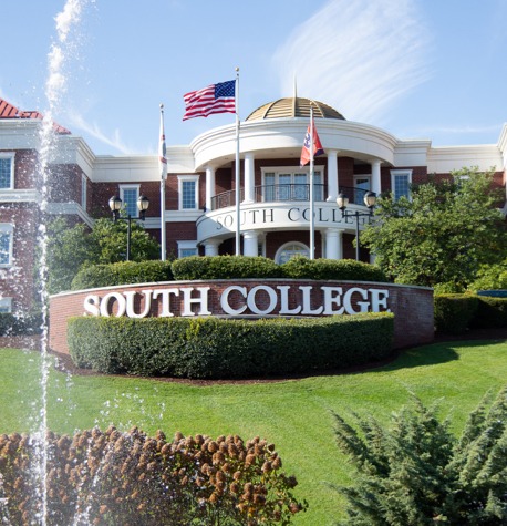 Exterior view of South College building with a large sign reading "South College" in front. The building is brick and features a dome. A fountain is visible in the foreground, and several flags, including the American flag, are flying above the entrance.