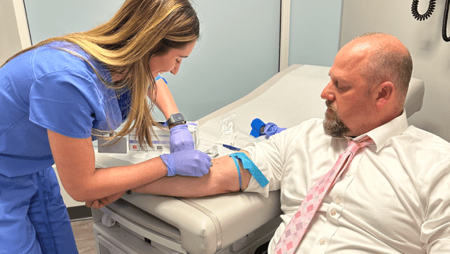 A medical assisting student wearing a blue uniform and gloves is drawing blood from a man sitting on a medical examination table. The man is dressed in a white shirt and pink tie.