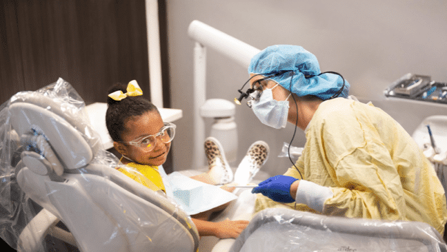 A child sits in a dental chair, wearing safety goggles and a yellow outfit with a matching bow. A dental student, dressed in protective gear including a face mask, gloves, and a hair cover, attends to them.