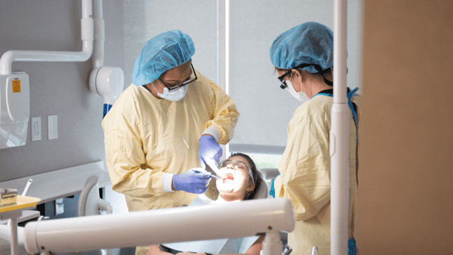Two dental students wearing protective gear are performing a dental procedure on a patient. One stands by the patient's head, using dental tools, while the other assists.