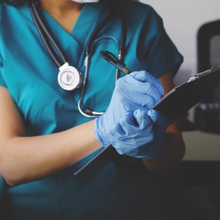 A nursing student wearing teal scrubs, blue gloves, and a stethoscope is writing on a clipboard with a pen.
