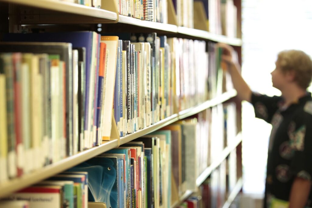 In a library aisle, a South College student is browsing through shelves filled with books. The focus remains on the colorful book spines, while the blurred figure reaches for a book in the background.