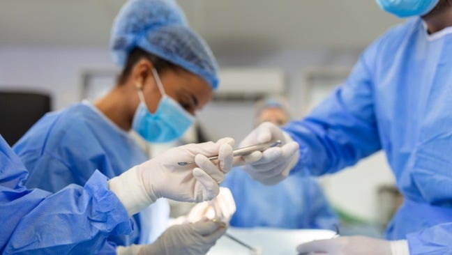 Students in blue scrubs and masks are in an operating room setting. One person hands a surgical instrument to another, highlighting teamwork.