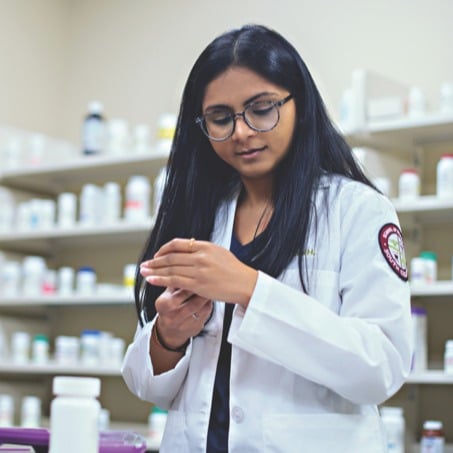 A pharmacy student with long black hair and glasses is wearing a white lab coat. She is standing in a laboratory with shelves stocked with various bottles in the background, focusing on something she is holding in her hands.