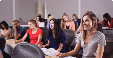 A diverse group of students in a classroom, sitting at desks with notebooks open. They appear attentive and engaged. The room has a neutral color scheme, with large windows in the background.