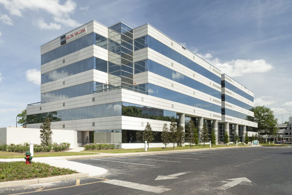 A modern, multi-story office building with glass windows reflecting the blue sky and white clouds. The structure is surrounded by landscaped greenery and features a company sign at the top corner. A paved parking lot and sidewalk are visible in the foreground.