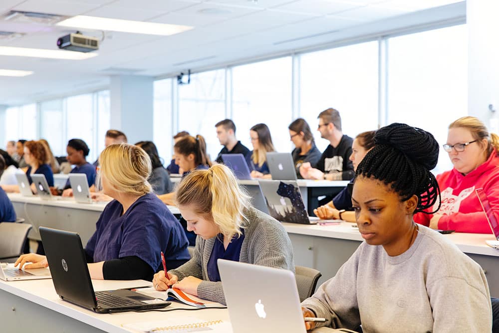 Students sitting in a brightly lit classroom, attentively using laptops and taking notes during a lecture.