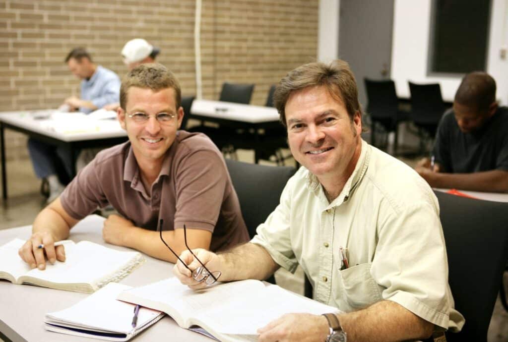 Two male students smiling at a table, each with an open book. The man on the right holds glasses. In the background, fellow students also read at desks in a classroom setting.