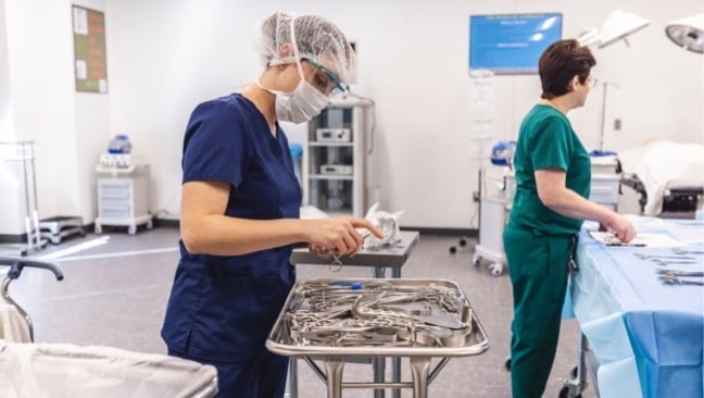 Two surgical technology students are in a hospital room. One in blue scrubs and a hairnet is inspecting and organizing surgical instruments on a tray. The other in green scrubs is preparing equipment near a patient bed.