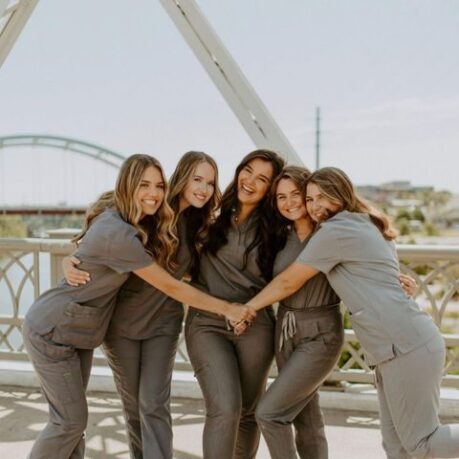 Five student dental assistants wearing gray scrubs stand on a bridge outdoors, hugging and smiling at the camera.