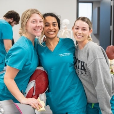 Three people smiling in a classroom. Two wearing teal scrubs and one in a gray sweatshirt.