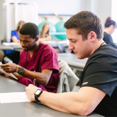 Two students sitting at a table in a classroom setting, focused on writing. One is wearing a black shirt and a smartwatch, while the other is in maroon scrubs. The background shows more people similarly engaged and some medical equipment.