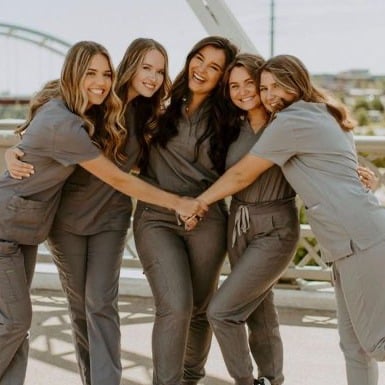 Five students wearing gray scrubs stand closely together, smiling and hugging on a sunny outdoor bridge. The background features a blue sky and a distant bridge, adding to the cheerful and supportive atmosphere of the group.