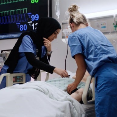 Two nursing students, one wearing a hijab, and the other with blonde hair in a bun, attend to a patient in a hospital bed. Medical monitors display vital signs in the background, showing heart rate, pulse, and blood pressure readings.