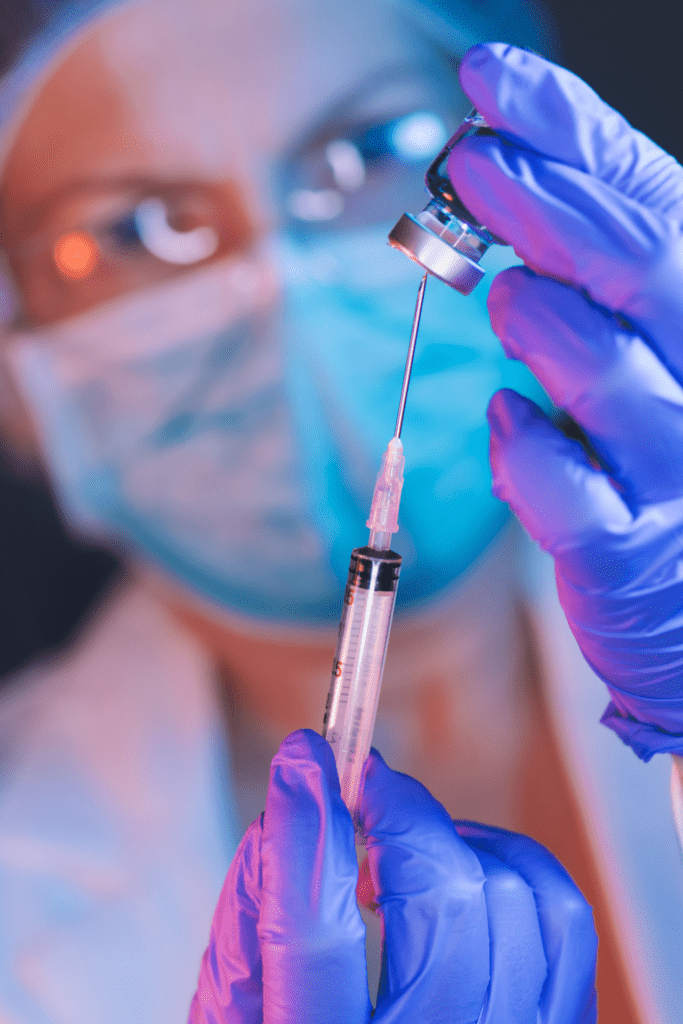 A student nurse anesthetist wearing a mask and gloves is preparing a syringe with a vaccine. The focus is on the needle and vial, with the person's face blurred in the background.