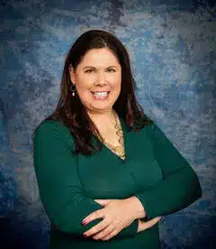 A woman with long dark hair is smiling and standing with her arms crossed against a textured blue background. She is wearing a green top and a necklace.