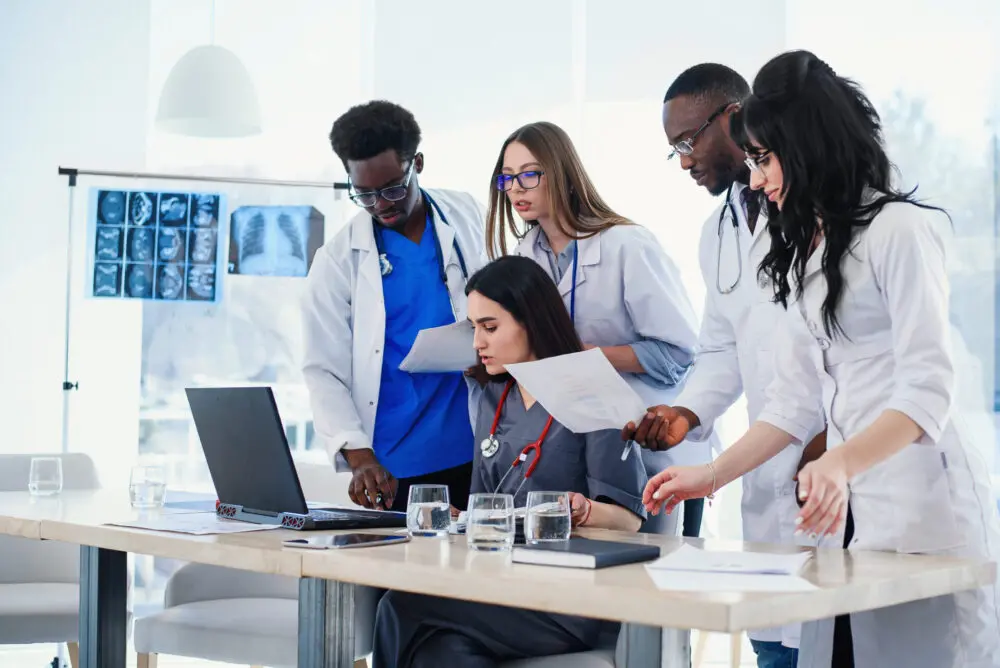 A diverse group of physician assistant students gather around a table, looking at documents and a laptop. Medical charts and X-rays are visible on a lightbox in the background. The team appears to be engaged in a discussion or collaborative review of medical information.