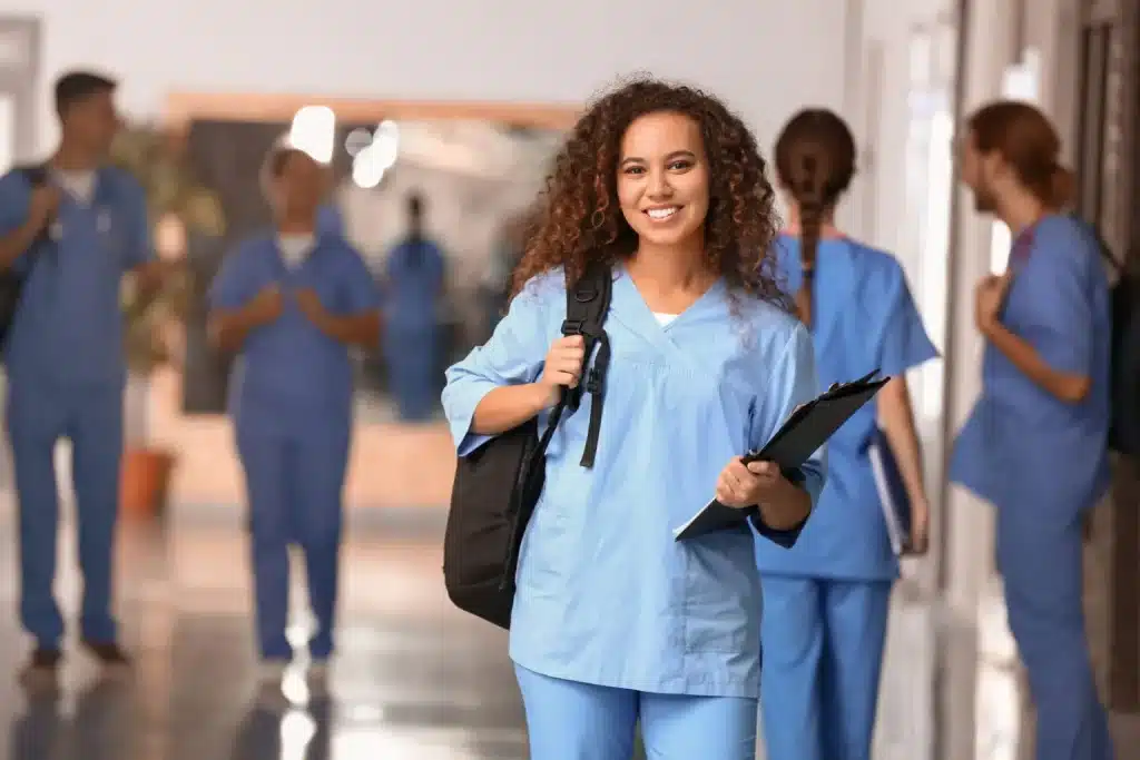 A smiling student dressed in light blue medical scrubs stands in the foreground, holding a clipboard and a backpack. In the background, several other individuals also in medical scrubs are walking and talking in a brightly lit hallway.