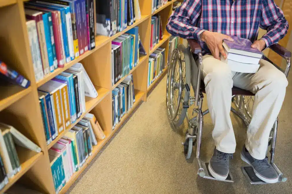 A person in a wheelchair holds multiple books while navigating through a library aisle. The bookshelves beside are filled with a variety of books.