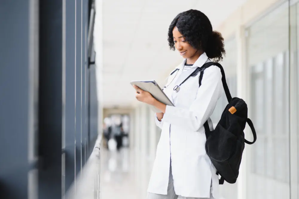 A female student wearing a white lab coat and stethoscope around her neck stands in a brightly lit hallway, looking at a tablet she is holding in her hands. She has a black backpack on one shoulder and appears to be smiling while focusing on the tablet.