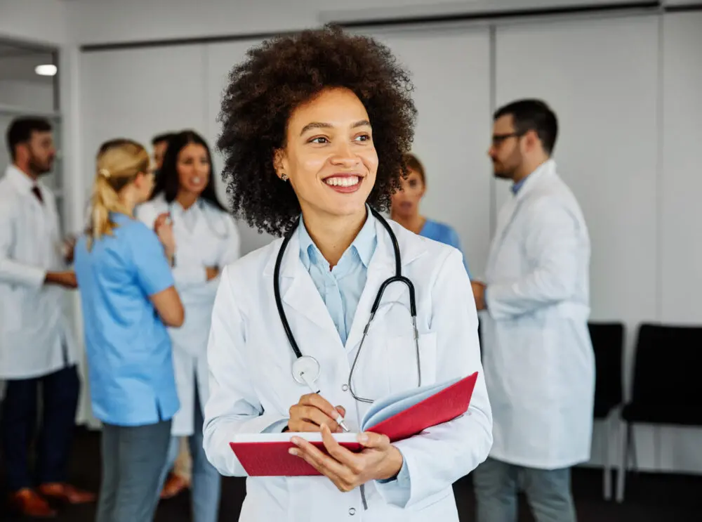 A smiling student physician assistant with a stethoscope around her neck holds a red notebook while standing in the foreground. In the background, a group of medical staff in white coats and scrubs engage in conversation. The setting appears to be a medical facility.