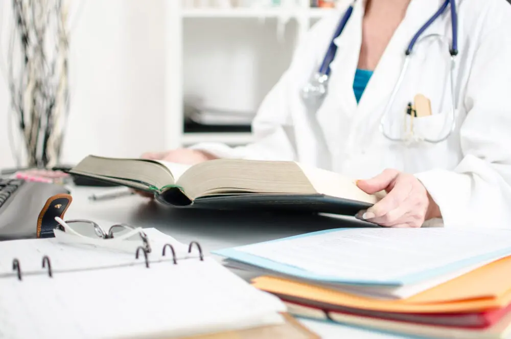 A person in a white lab coat is seated at a desk reading an open book. On the desk, there are various documents, a binder, a pen, and a stethoscope.