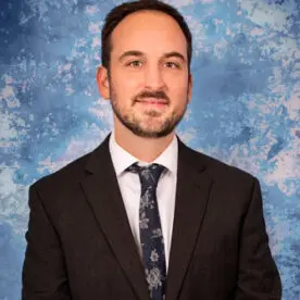 A man with short dark hair and a beard, wearing a suit, white shirt, and patterned tie, stands against a blue and white marbled background. He is looking directly at the camera with a slight smile.