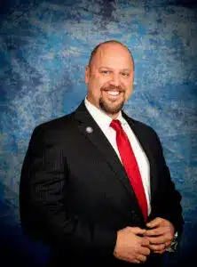 A smiling man is dressed in a black suit with a white shirt and a red tie, standing against a blue, textured background. He has a short, neatly trimmed beard and is posing with his hands clasped in front.