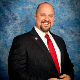 A man dressed in a dark suit with a white shirt and a bright red tie smiles at the camera. He is standing against a blue and white marbled background. He stands with his hands clasped in front of him.
