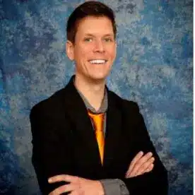 A person with short brown hair is smiling with arms crossed in front of a blue marbled backdrop. They are wearing a black suit jacket, a grey shirt, and an orange tie.