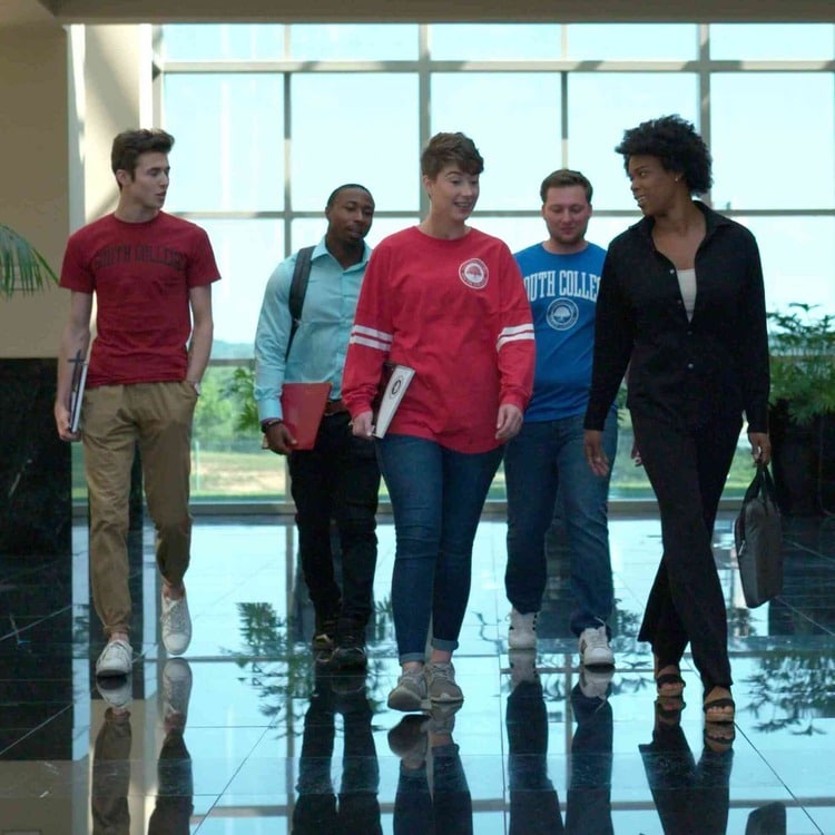 Five students walk through a brightly lit hallway with large windows. They are engaged in conversation and appear to be friends. Two of them hold folders. They are casually dressed, with some wearing college-branded shirts. The background shows greenery outside.