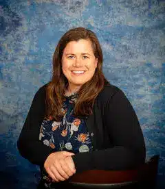 A person with long brown hair, wearing a floral blouse and black cardigan, smiles while sitting and resting their arms on a chair against a blue textured background.