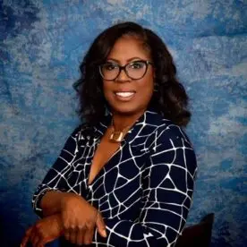 A woman with shoulder-length black hair and glasses stands against a blue textured background. She is wearing a dark blue dress with white geometric patterns and a gold necklace, and she is smiling while looking at the camera. Her arms are crossed in front of her.