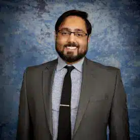 A man with glasses and a neatly trimmed beard smiles at the camera. He is wearing a gray suit with a striped shirt and a black tie. The background is a mottled blue texture.