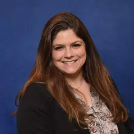 A woman with long brown hair is smiling and looking at the camera. She is wearing a floral-patterned blouse under a dark blazer. The background is a solid blue color.