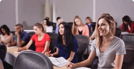 A classroom filled with students sitting at desks. Most of the students are focused on their work, while a few are looking up and listening. One female student in the foreground is smiling and appears engaged in a discussion. Each student has notebooks and pens.