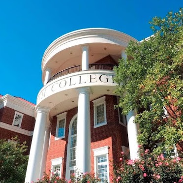 A red brick building with large white columns at the entrance, adorned with an arch that has partially visible text, hinting at it being a college. The facade is surrounded by greenery and colorful flowers under a clear blue sky.