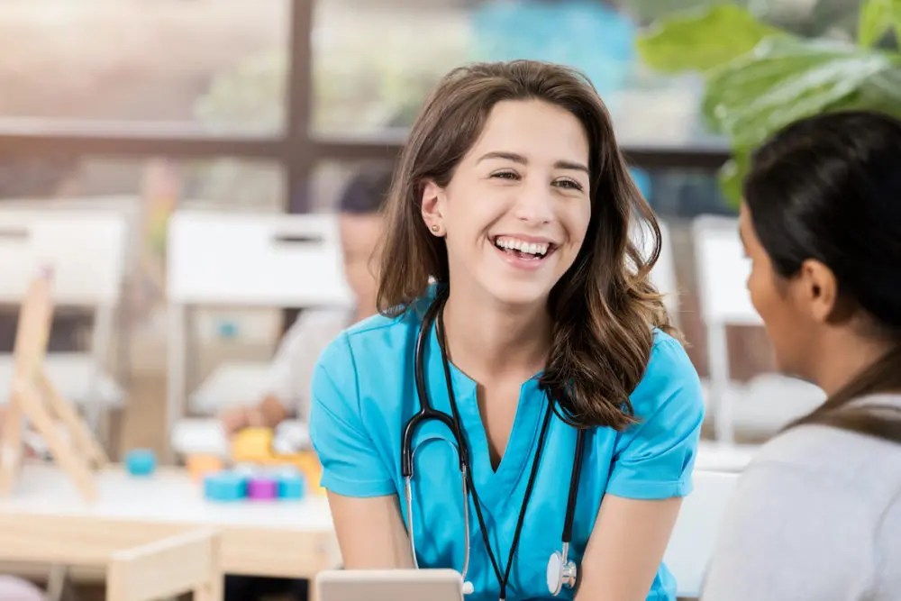 A female student physician assistant in scrubs, with a stethoscope around her neck, is smiling and talking to another person, who is partially visible. The background shows a well-lit room with desks and plants.
