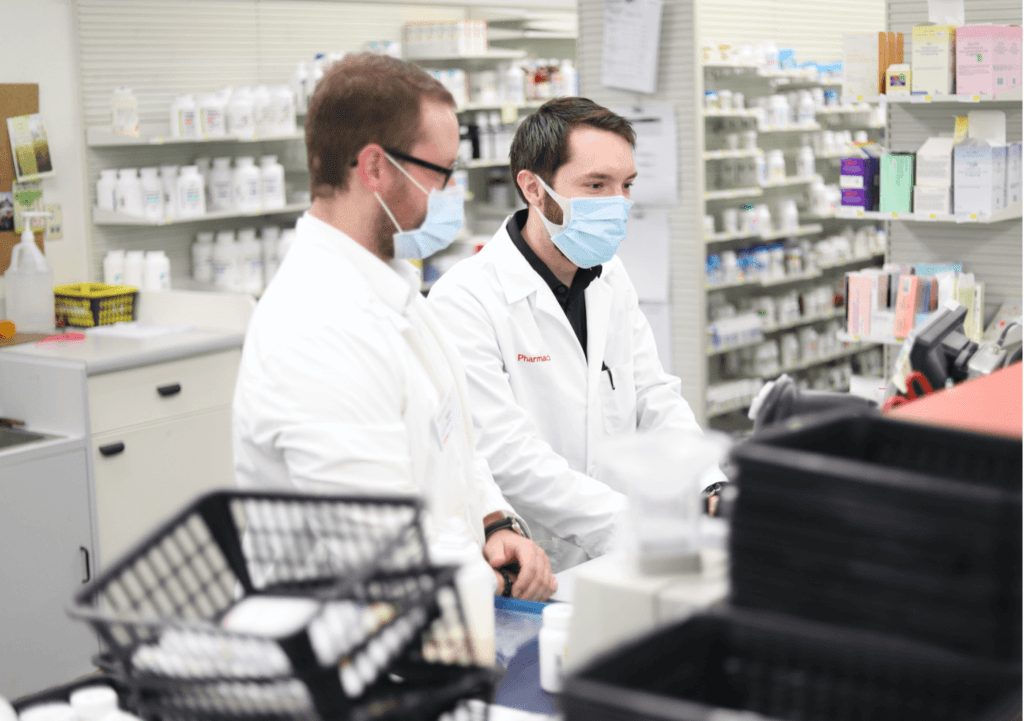 Two pharmacy students wearing white lab coats and face masks are behind a pharmacy counter, working on a computer. Shelves filled with various medications and pharmaceutical products are visible in the background.