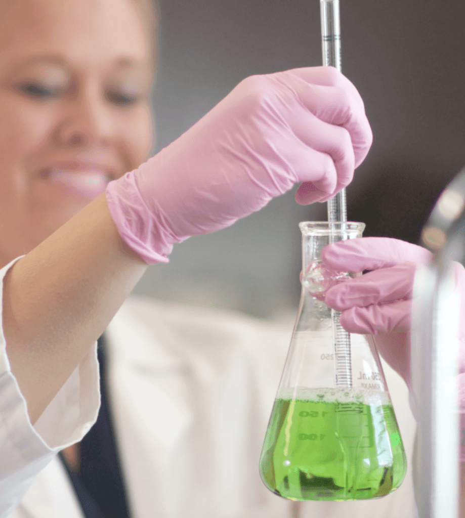 A student wearing pink gloves is holding a glass pipette and a conical flask containing green liquid. The person is dressed in a white lab coat.