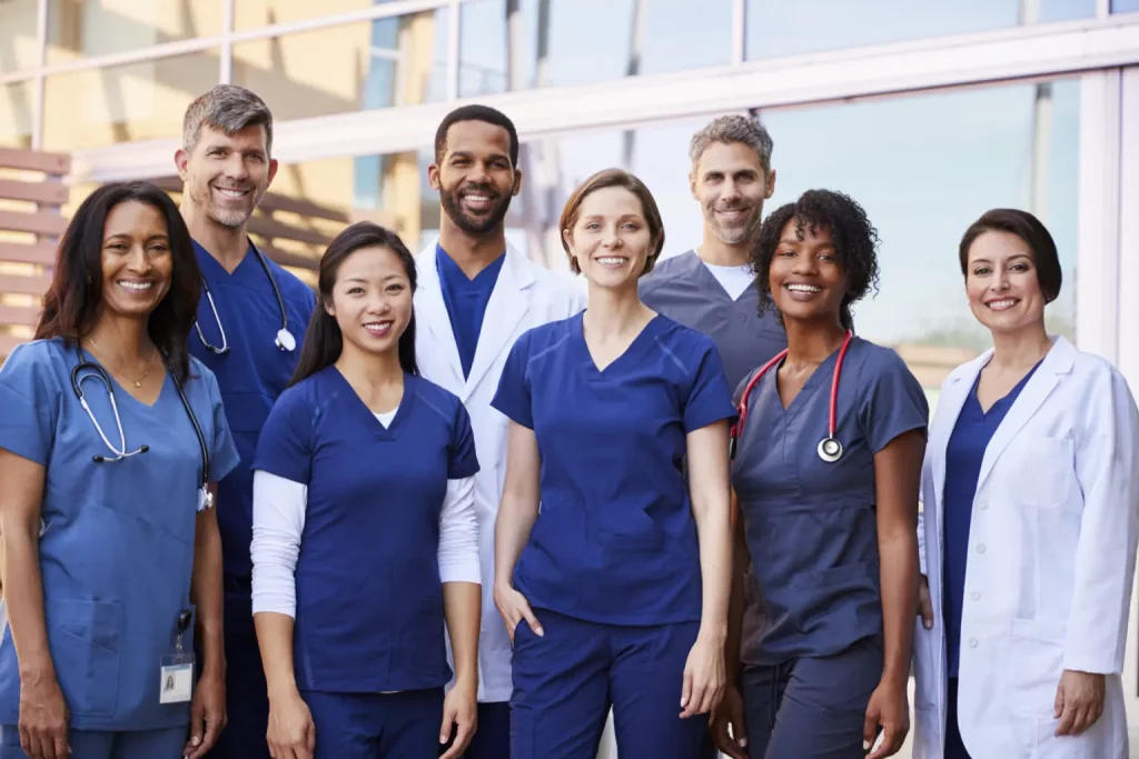 A diverse group of Physician Assistant students, including men and women, are standing together outside a building. They are wearing a mix of medical scrubs and white coats. Most are smiling and looking at the camera. The background features windows and part of the building.