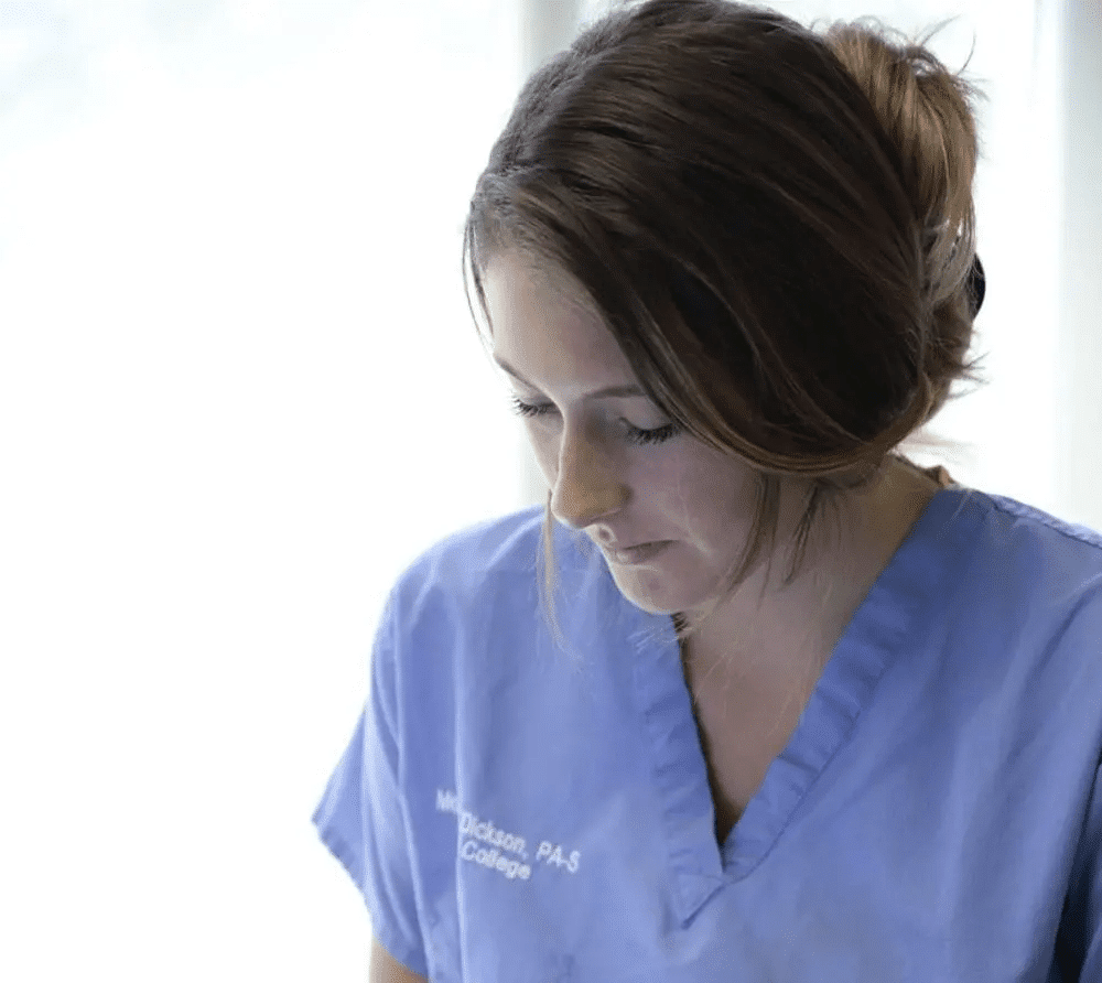 A student physician assistant with brown hair tied back is wearing blue medical scrubs and looking downward. The sunlight streams in from a window behind them, softly illuminating their face. The text on the scrubs reads, "PA-S South College.