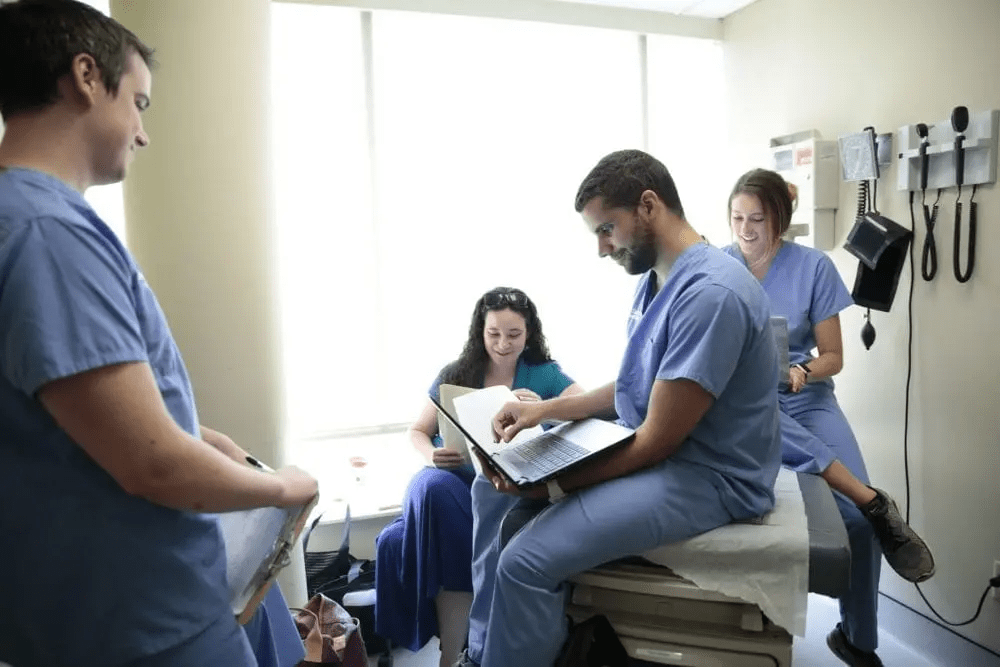 A group of four physician assistant students in blue scrubs are gathered in a small clinical room. Three are using laptops and paperwork, while one is sitting on an examination table. Medical equipment is hanging on the wall in the background.