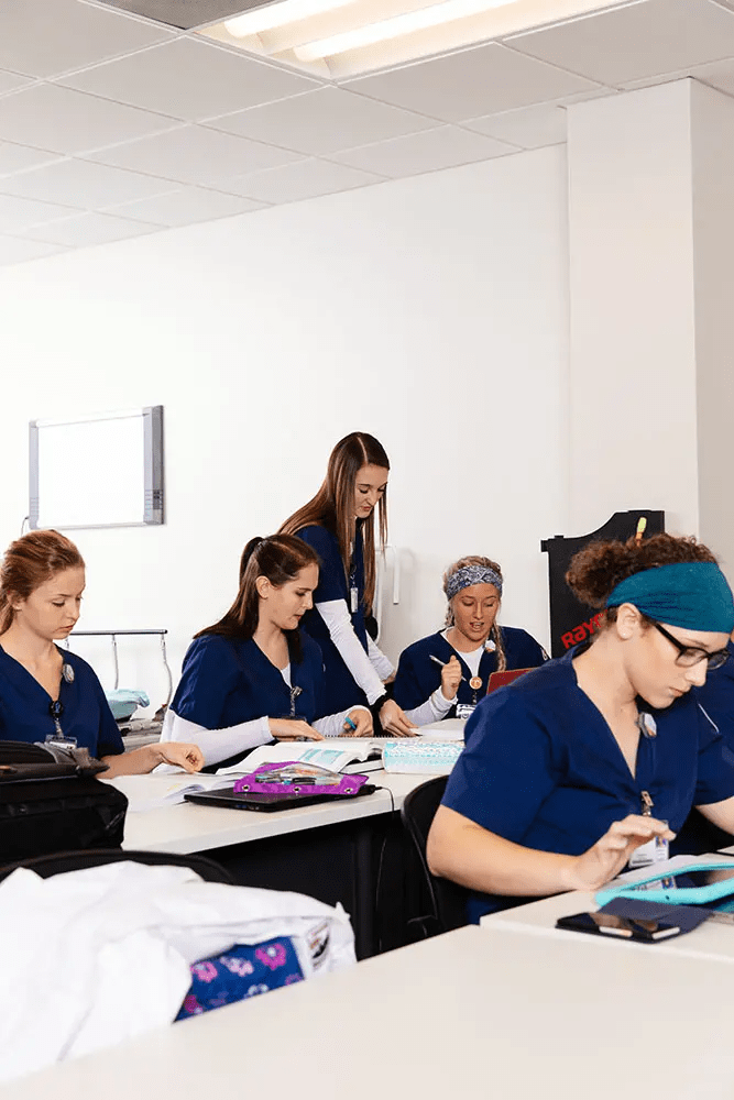 A group of nursing students in blue scrubs are working together at tables in a classroom. Some students are writing, while others are looking at papers or using tablets. A female instructor stands and assists a student in the middle. The atmosphere is focused and collaborative.