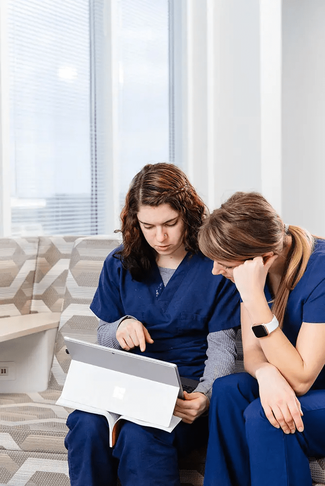 Two students in blue scrubs are seated on patterned chairs, focusing on a tablet. One is pointing at the screen, and the other is looking on thoughtfully.